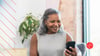 Woman around 65 wearing a grey shirt, playing games on phone and smiling. There is a plant in the background, and she is sitting on a couch.